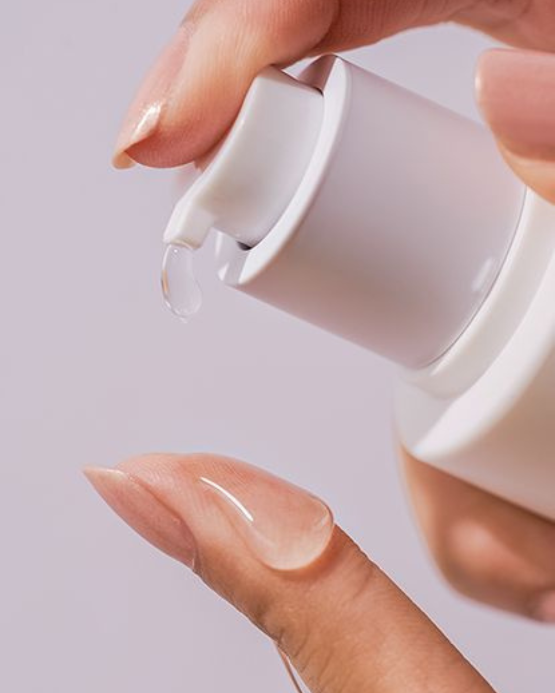 Hand holding a white bottle with a pump, dispensing a clear liquid onto a finger against a light purple background.